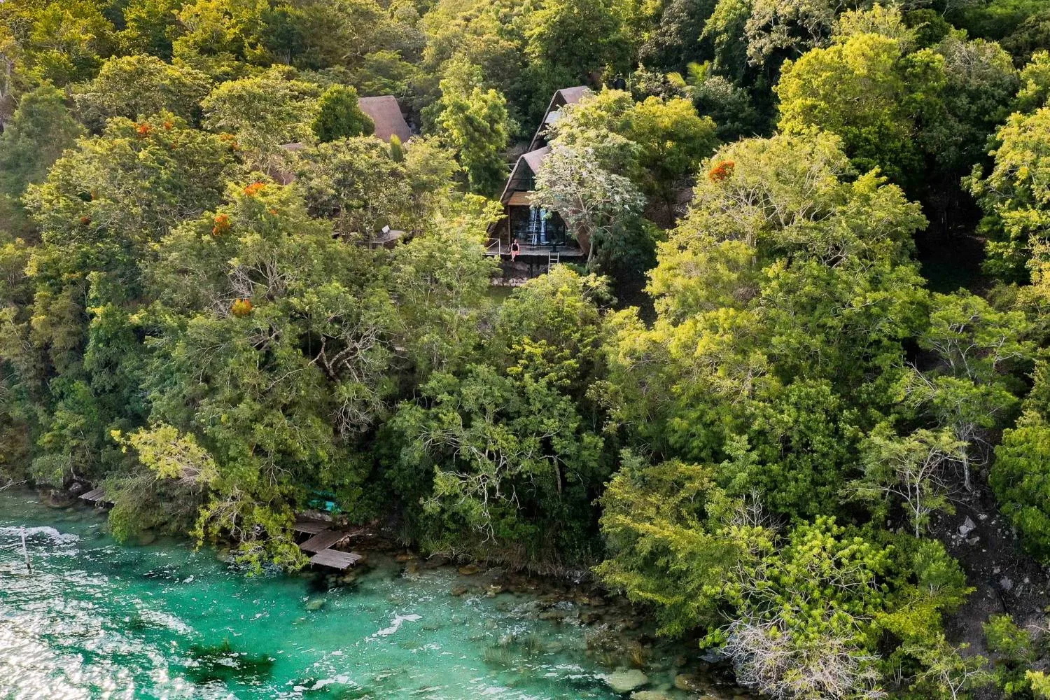Vista aérea de cabañas en forma de A entre la selva, sobre la orilla de la laguna de Bacalar, con un muelle de madera y el agua turquesa