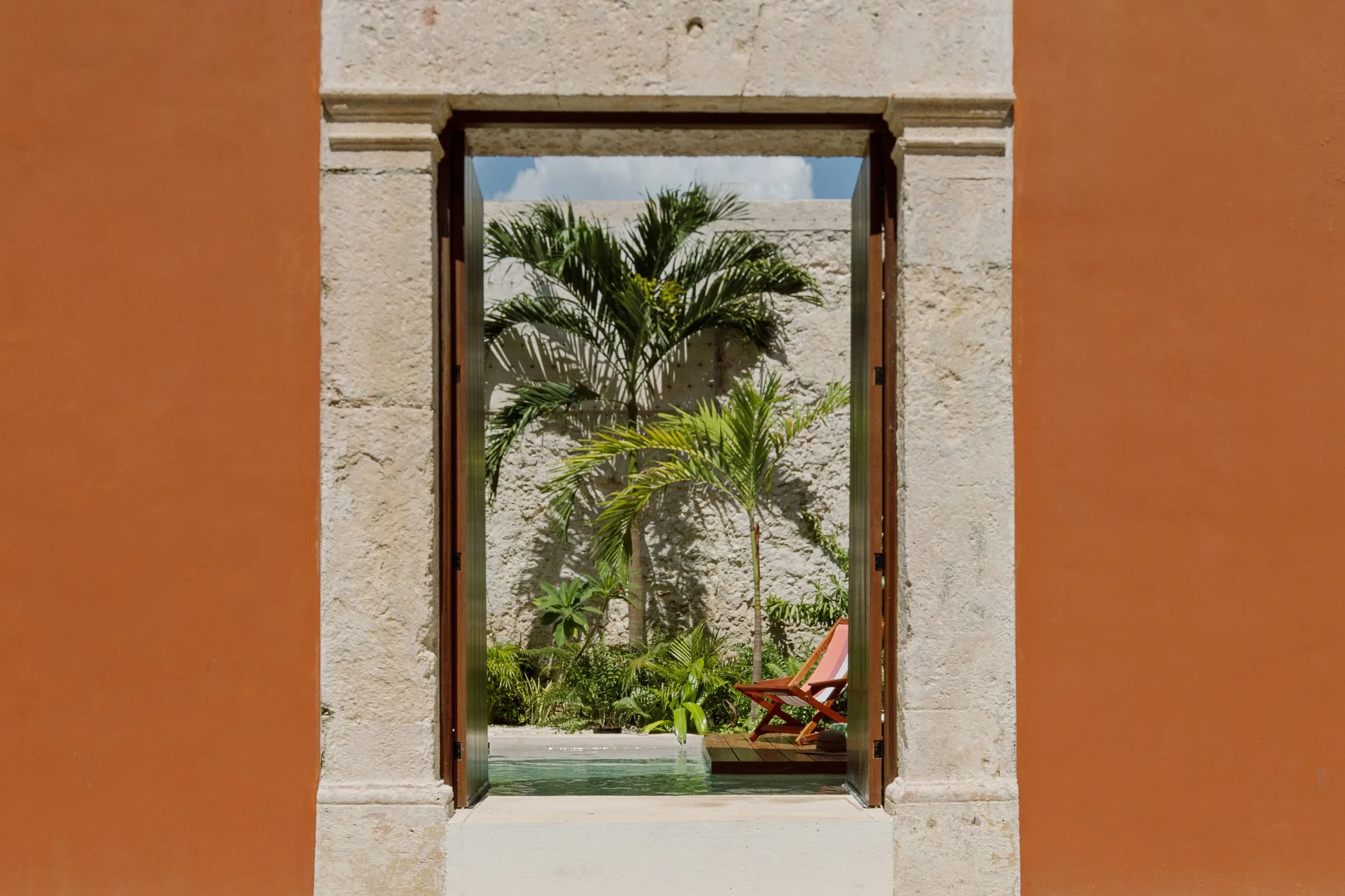 Puerta abierta en la fachada terracota de Casa Ex Templo, con palmeras y la alberca del patio al fondo, en la ciudad amurallada de Campeche. Casonas MX