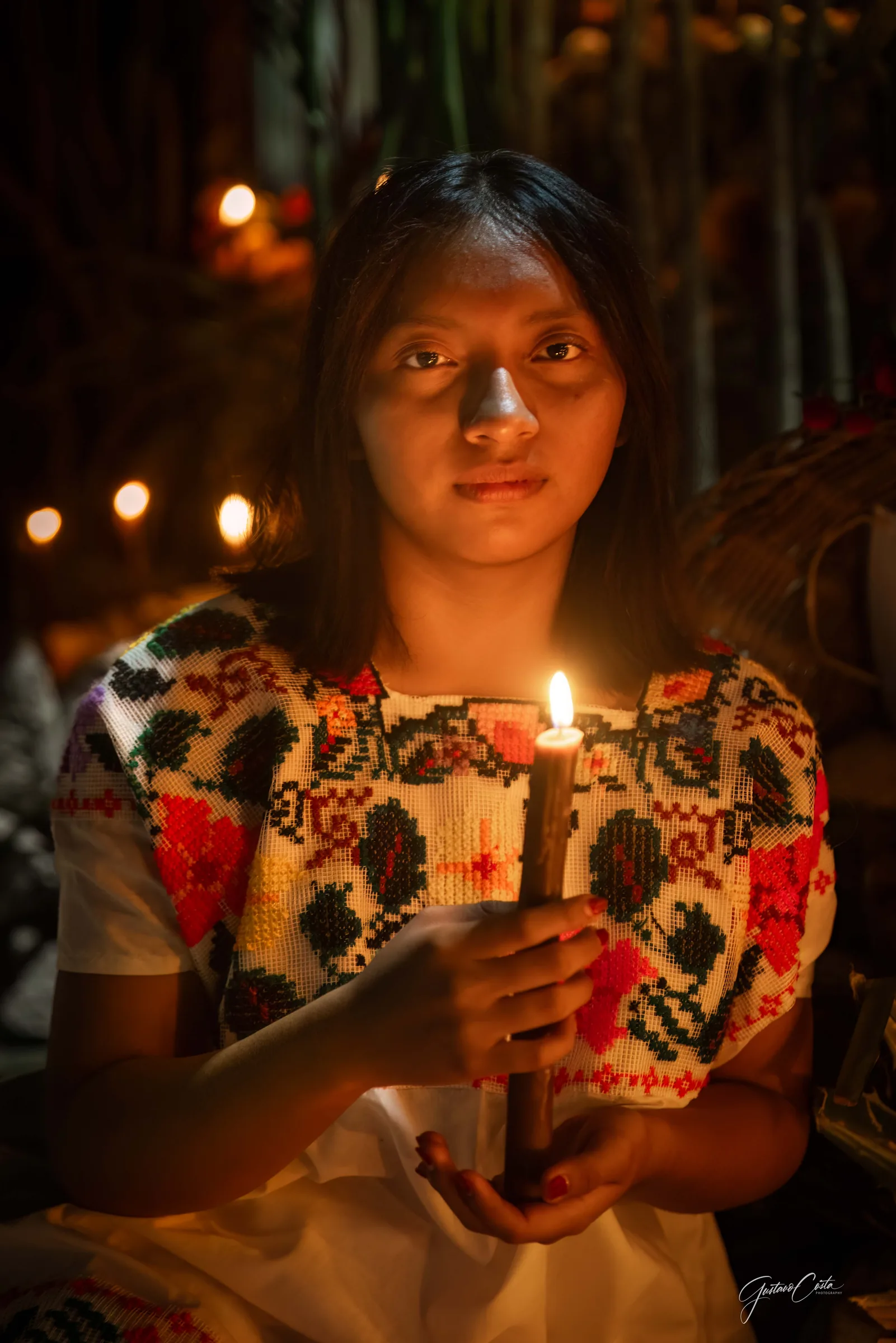 Joven con huipil bordado sostiene una vela encendida frente a un altar de Hanal Pixán