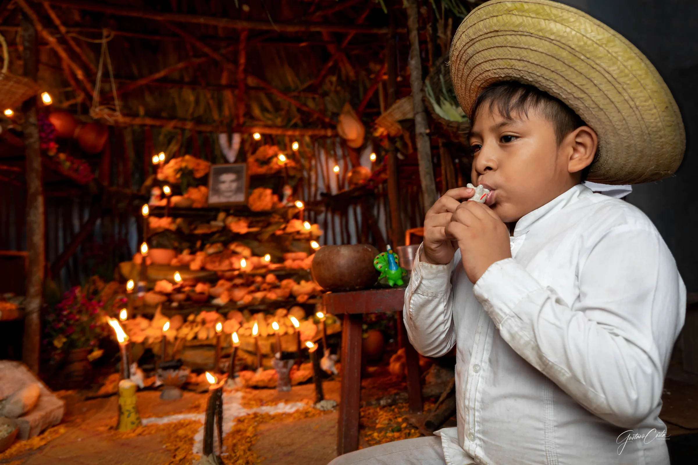 Niño con sombrero de palma toca un silbato de barro junto a un altar de Hanal Pixán iluminado con velas