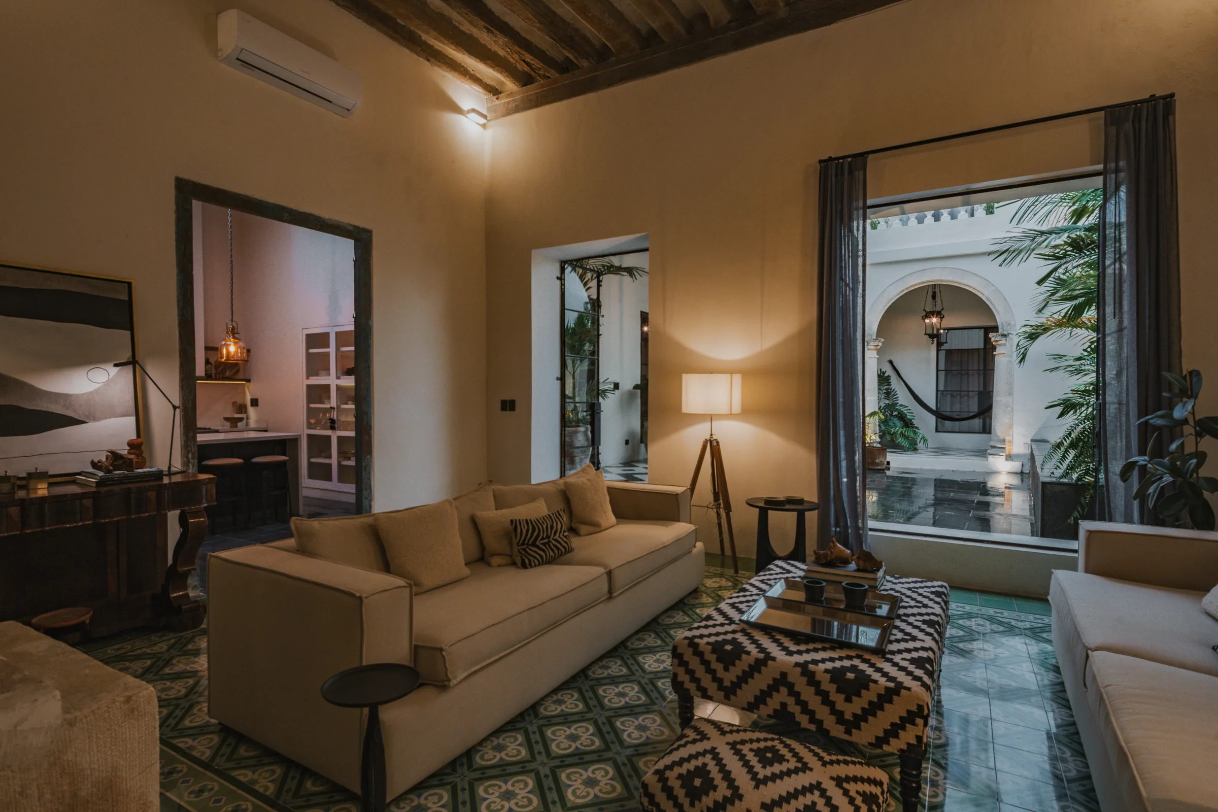 Sala de Casa Japa con vigas de madera y piso de pasta, abierta a un patio con arcos, palmeras y una hamaca
